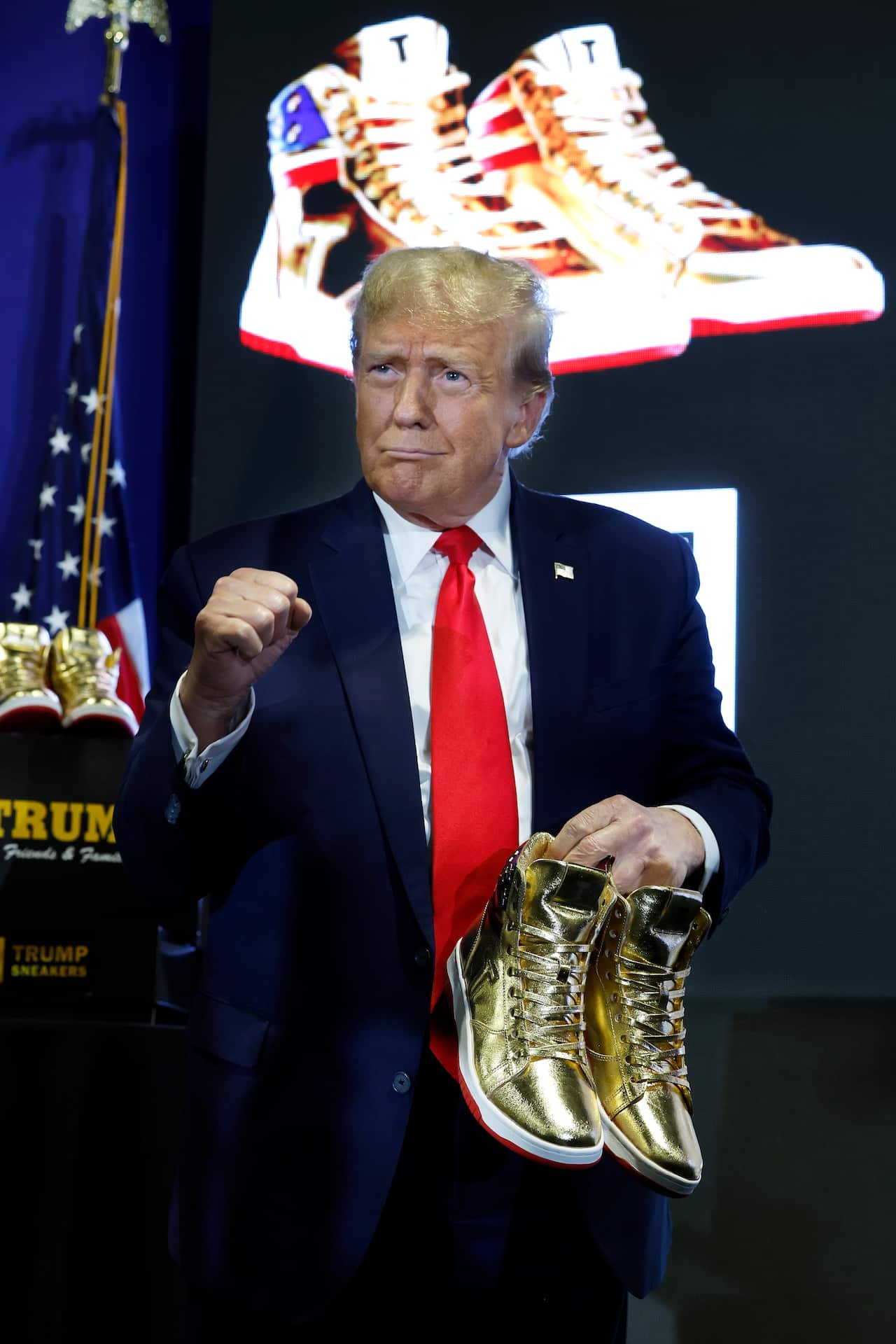 A man wearing a suit and tie holds a pair of gold-coloured shoes while standing on a stage.