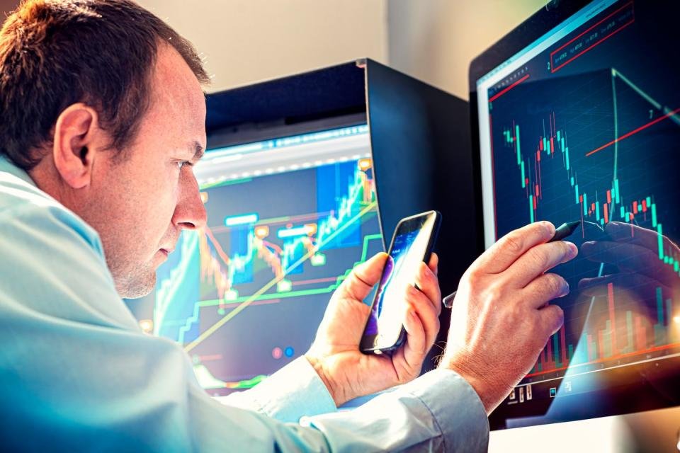 A professional money manager using a stylus and smartphone to analyze a stock chart displayed on a computer screen.