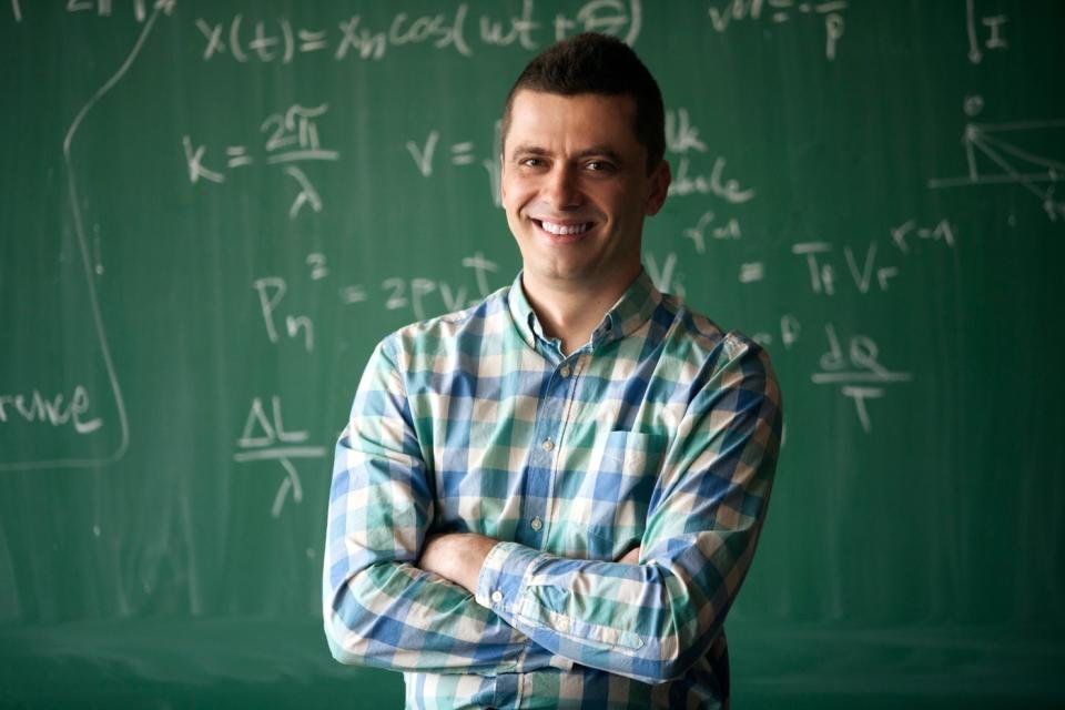 Someone with arms folded is smiling in front of a blackboard.