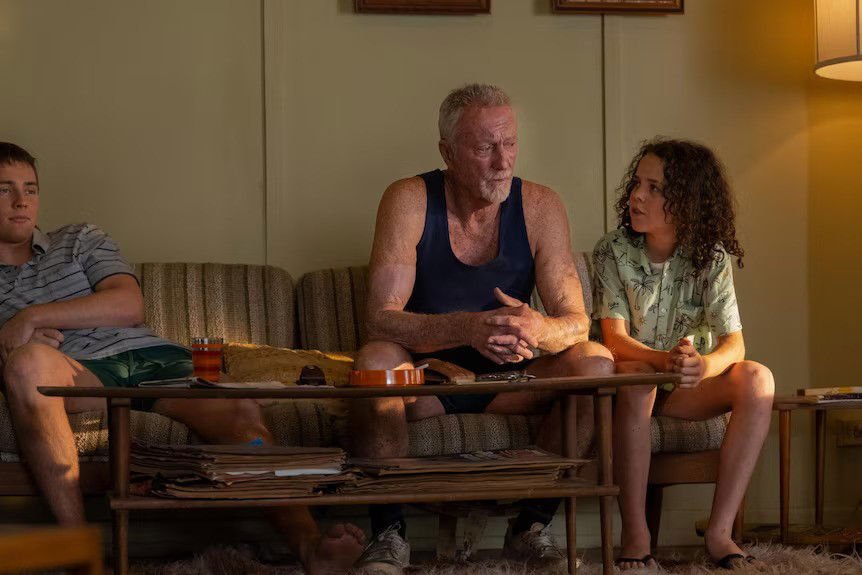 A TV still of Lee Tiger Halley, Bryan Brown and Felix Cameron sitting on a 70s-style couch.