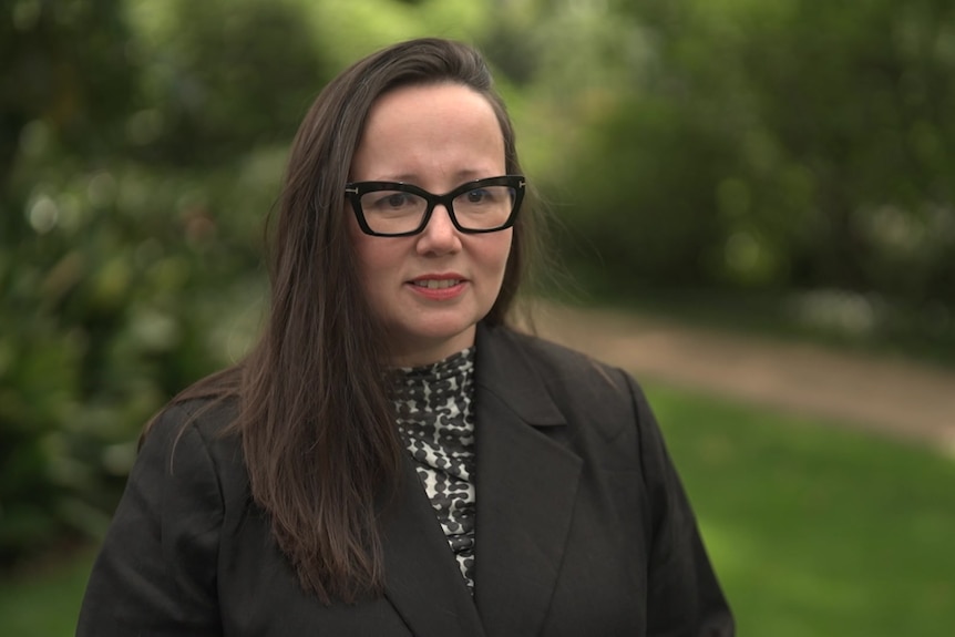 A woman earing glasses stands in a garden and looks at a camera