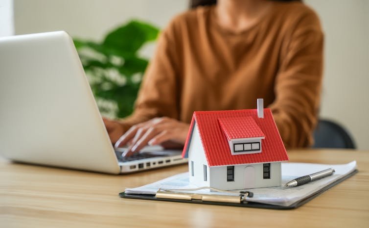 Woman on laptop with a clipboard and small model house in foreground